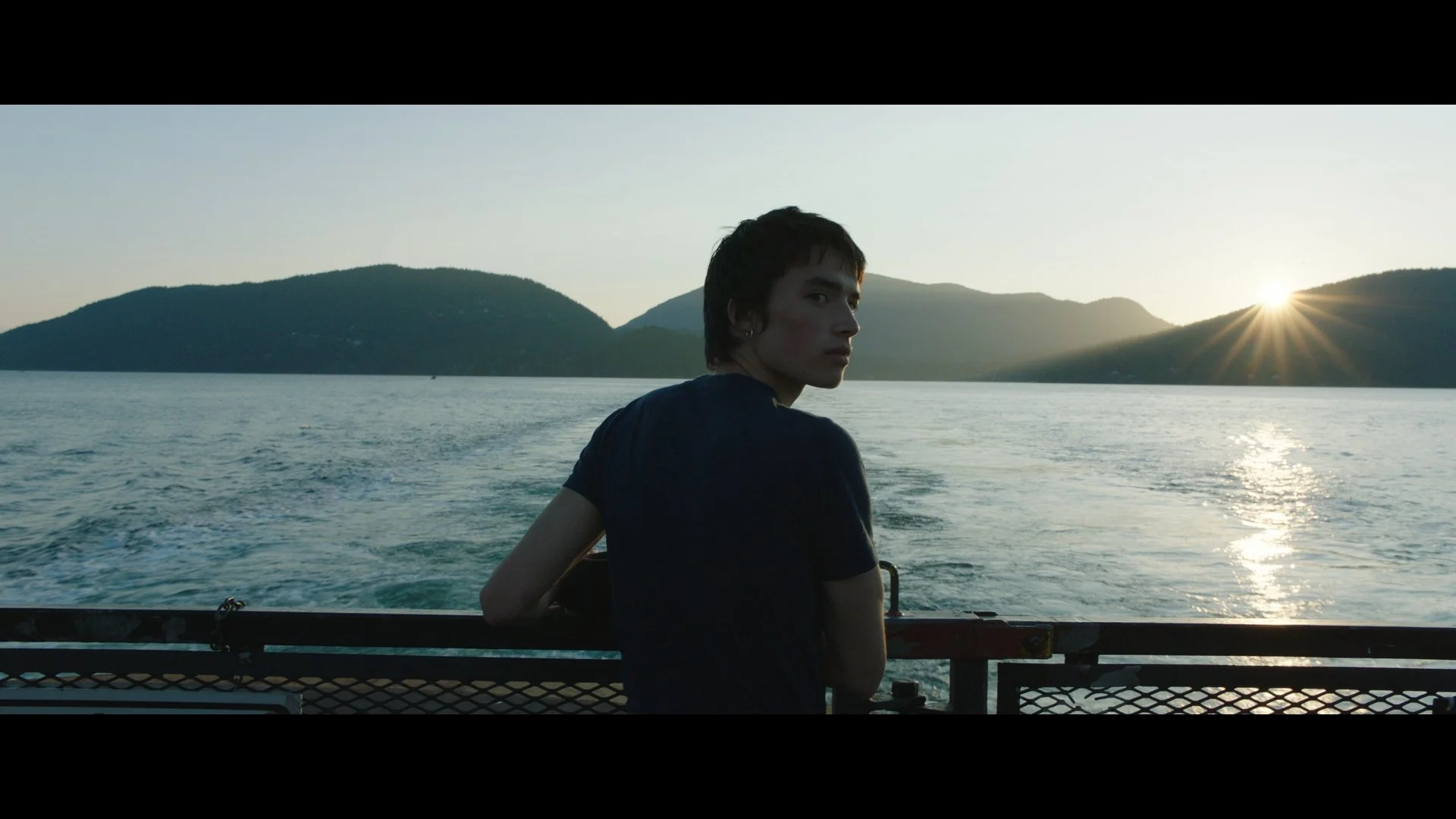 Film still from Manhunt: a young man at the stern of a ferry looking back toward the camera at sunset, mountains and water behind him.
