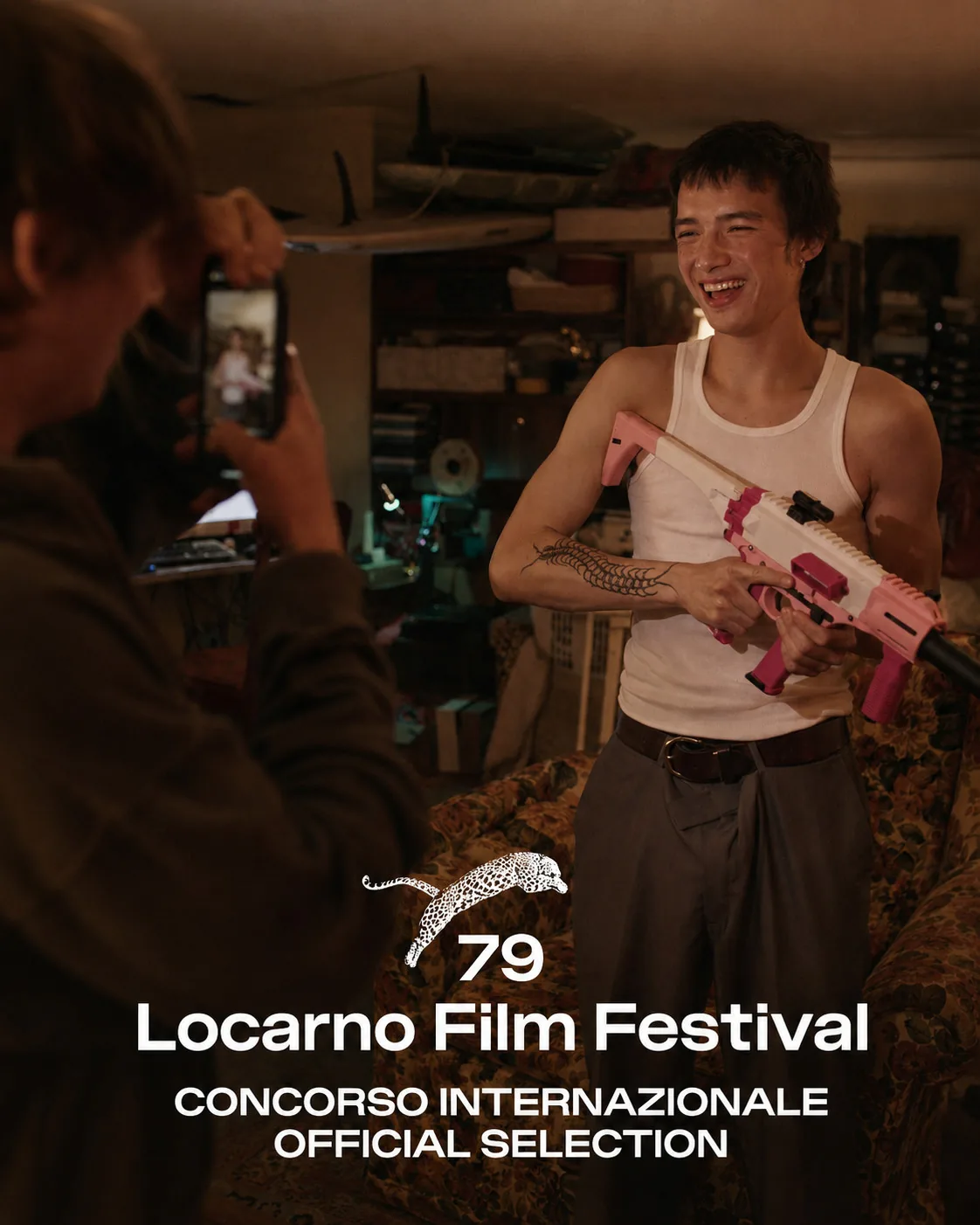 Film still from Manhunt beneath the 79th Locarno Film Festival Concorso Internazionale Official Selection laurel: a grinning young man in a white tank top holds a pink rifle in a cluttered room while another figure photographs him on a phone.