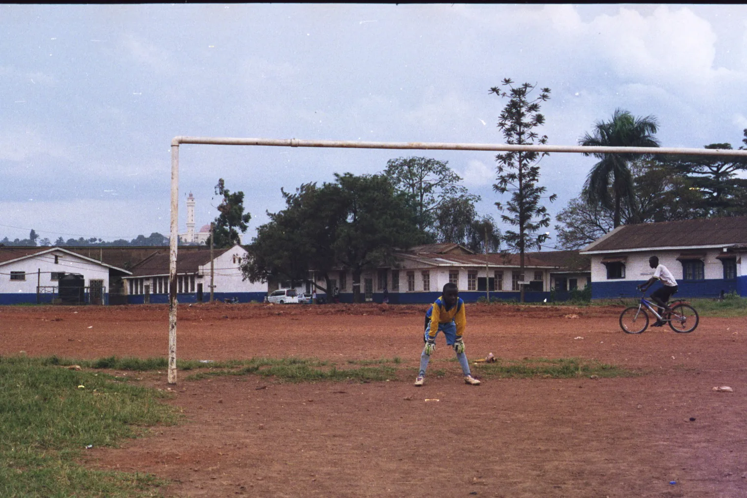 A young goalkeeper in yellow crouches by a weathered goalpost on a dirt football field while a boy rides a bicycle past low buildings.