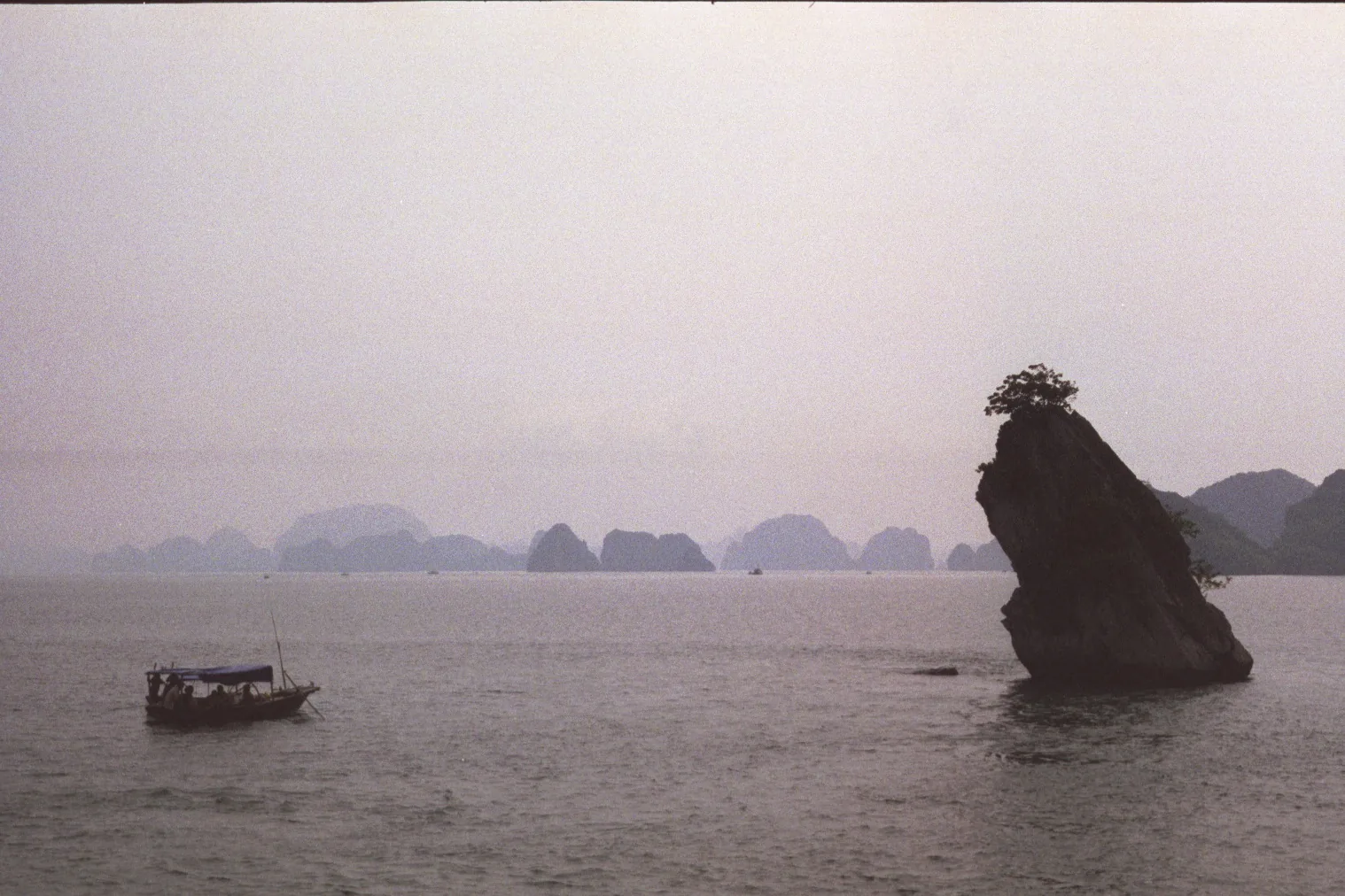 Small wooden fishing boat drifts past a jagged limestone outcrop on calm hazy water, with misty karst peaks beyond.