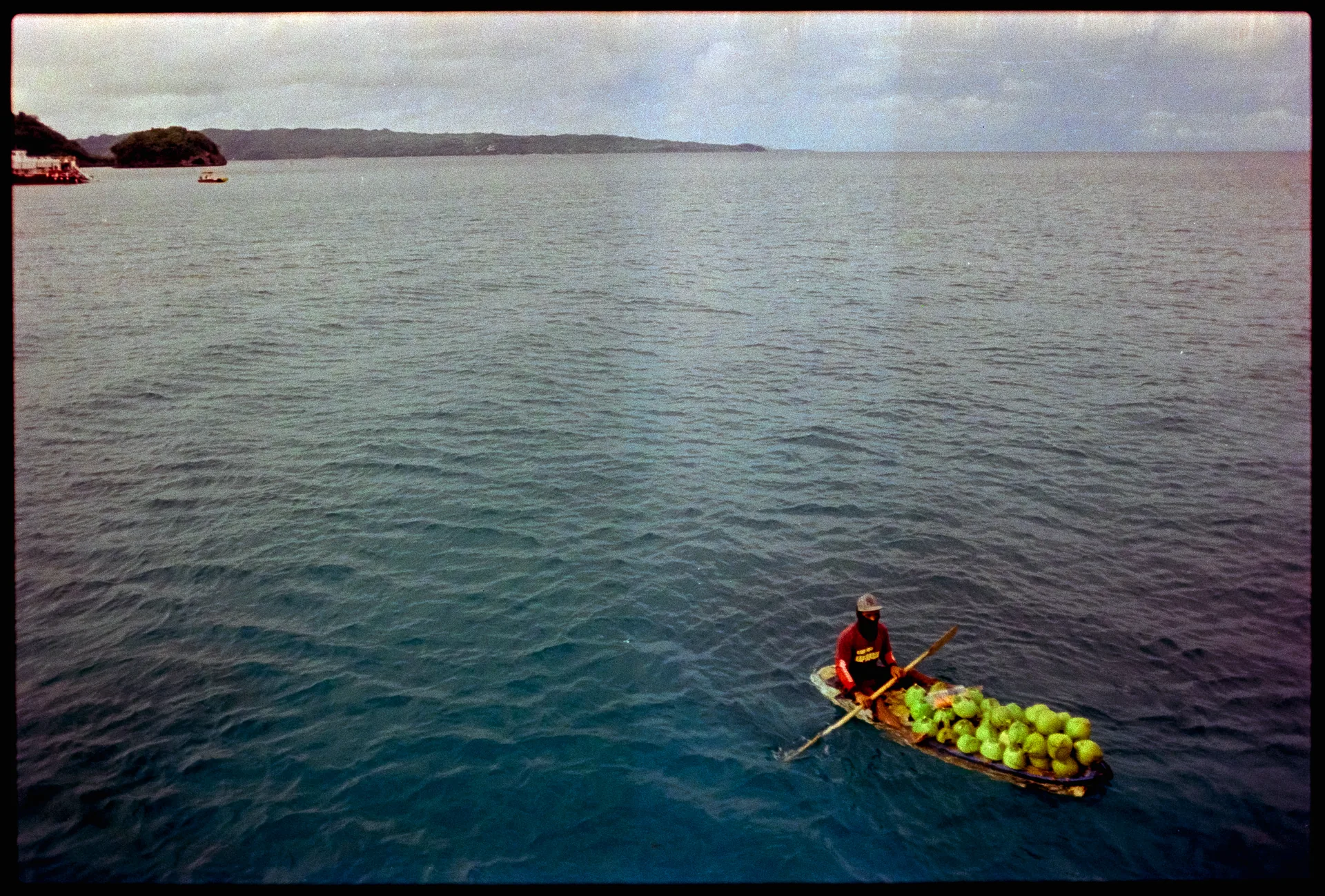 A person in an orange shirt paddles a slender wooden boat piled with green coconuts across deep blue open water near a distant shore.