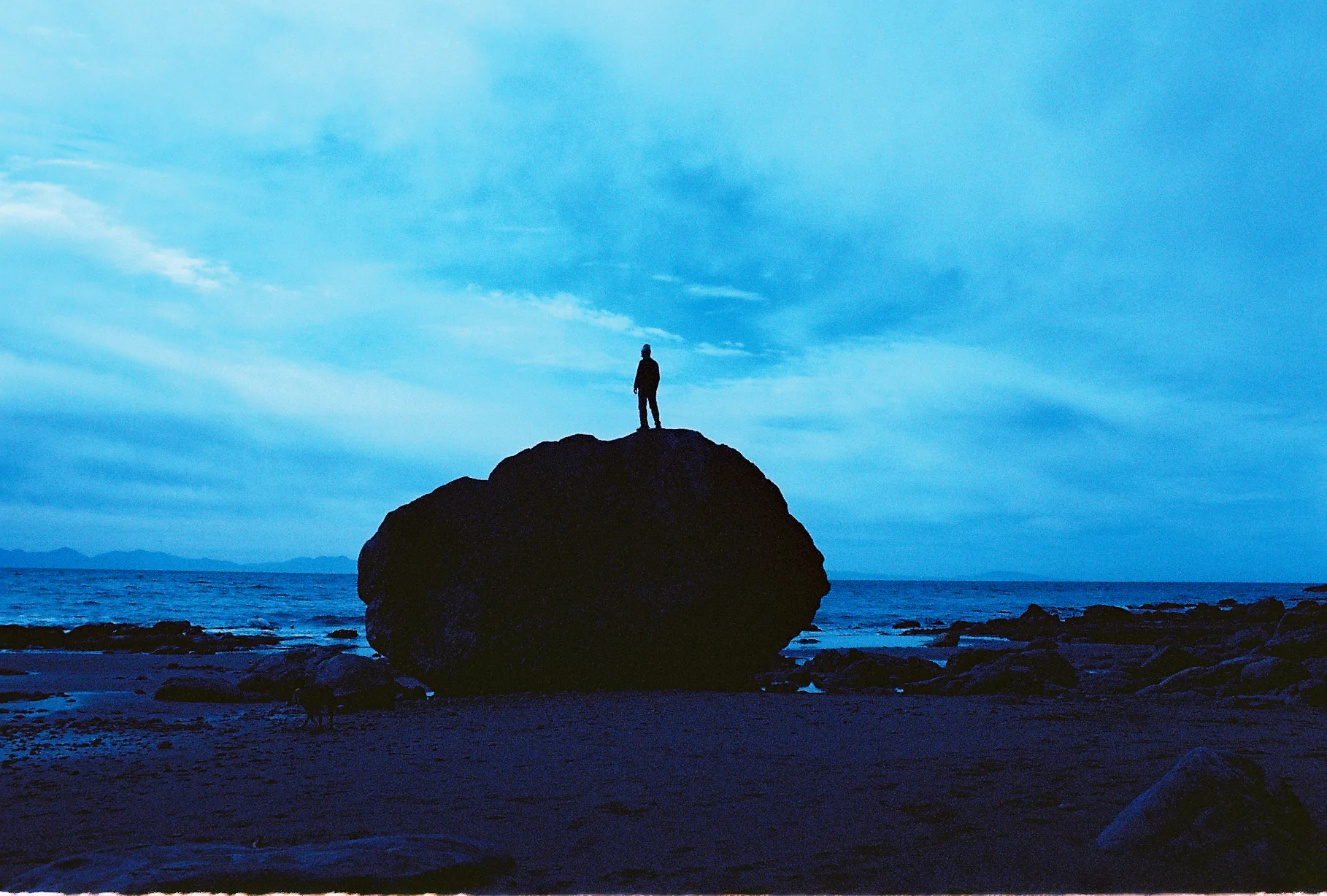 A person stands silhouetted on a large rounded boulder on a rocky beach against deep blue twilight sky and calm sea.