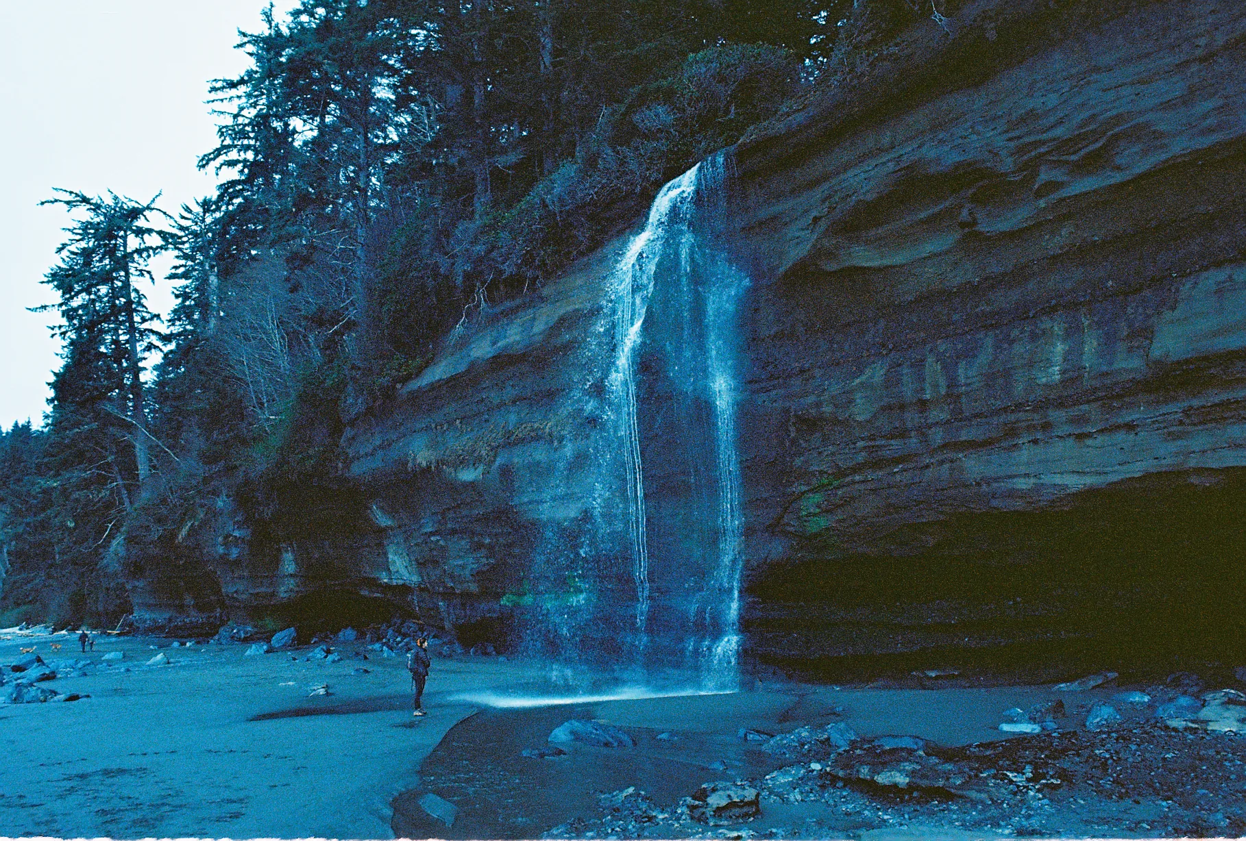 A thin waterfall spills down a layered sandstone sea cliff onto a rocky beach, with a small person standing at its base near the trees.