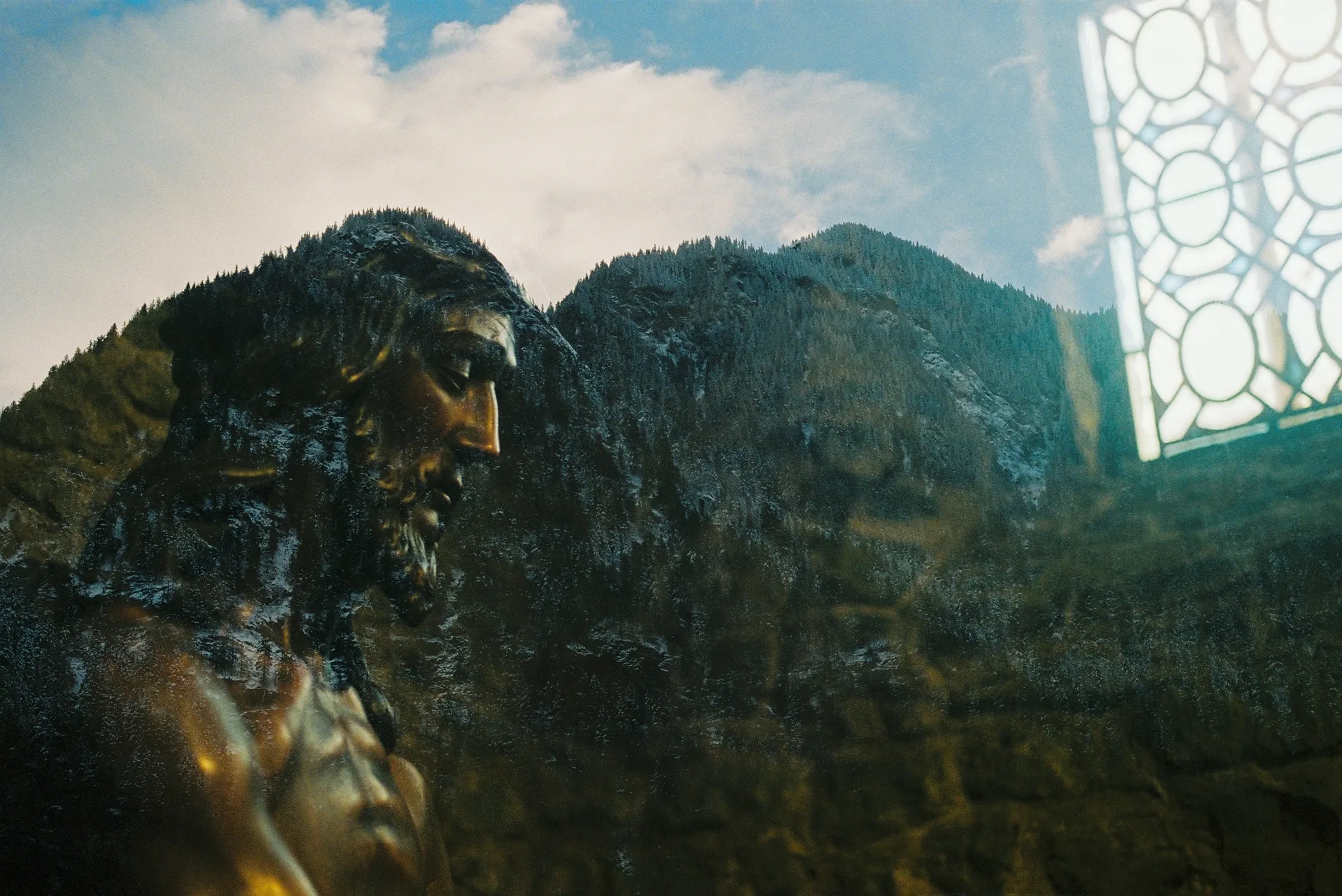 Double exposure blending a bronze sculpture of Christ in profile with a forested mountain ridge and patterned window light under a cloudy sky.