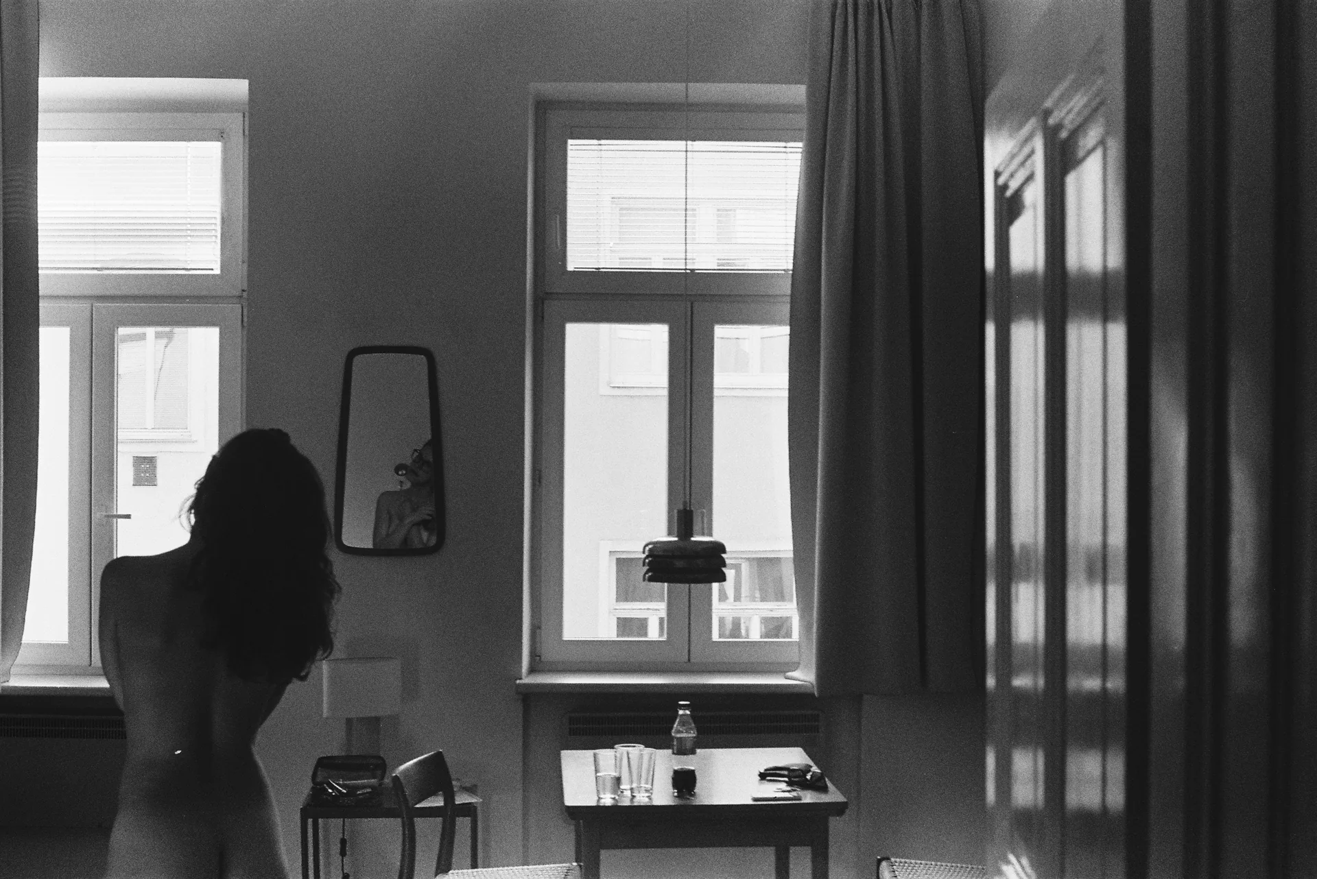 Black and white interior where a nude woman stands silhouetted before tall windows overlooking buildings, with a mirror and dressing table nearby.