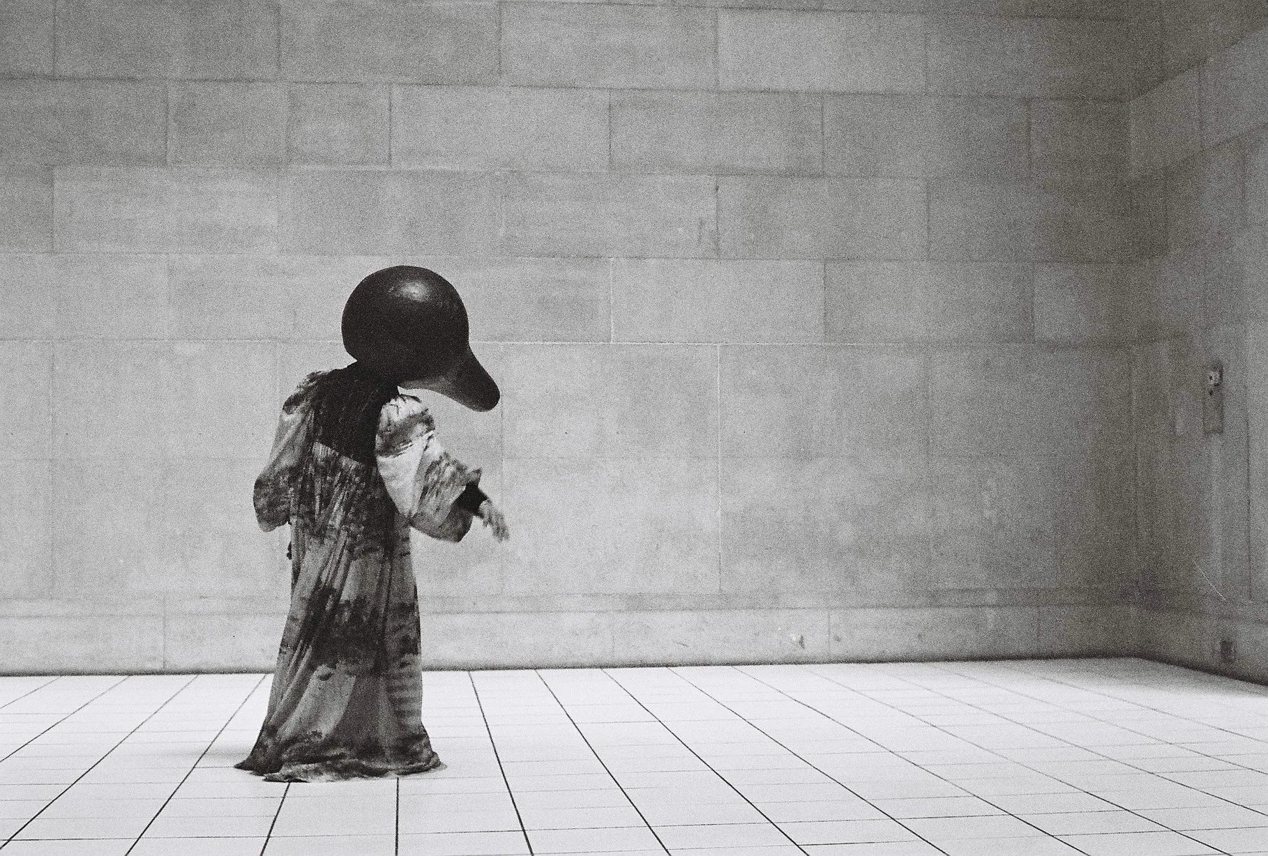 Black and white image of a small stooped figure wearing a dark domed helmet and tattered robe standing alone in an empty tiled room.