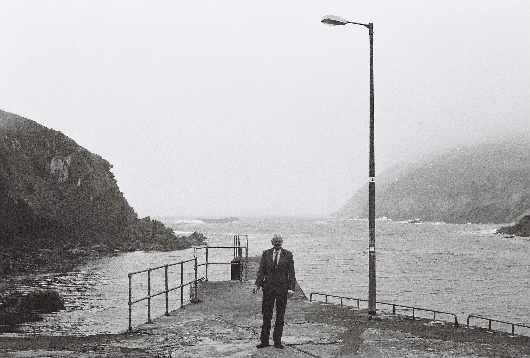 Black and white photo of a man in a suit and cap standing on a concrete pier beside a lamppost, with foggy cliffs and rough sea behind.