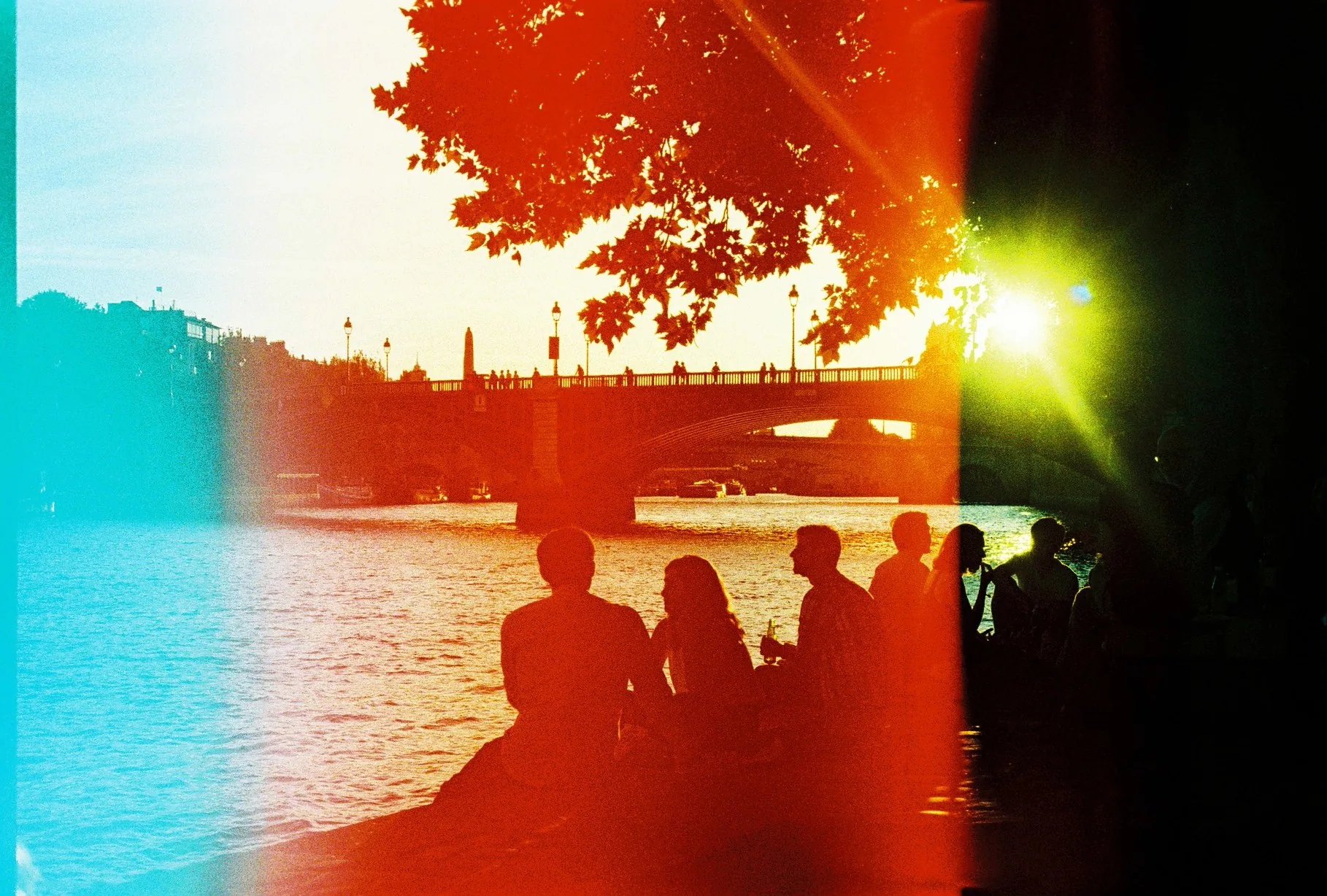 Silhouetted group sits on a riverbank as the sun sets behind a Seine bridge, with heavy red and teal film light leaks across the frame.