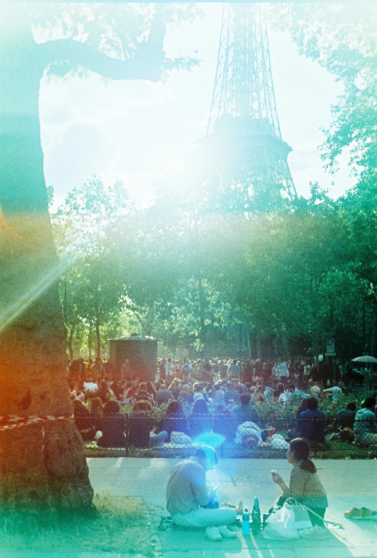 People picnic on grass beneath trees with the Eiffel Tower rising in the background, overlaid by green and orange film light leaks.