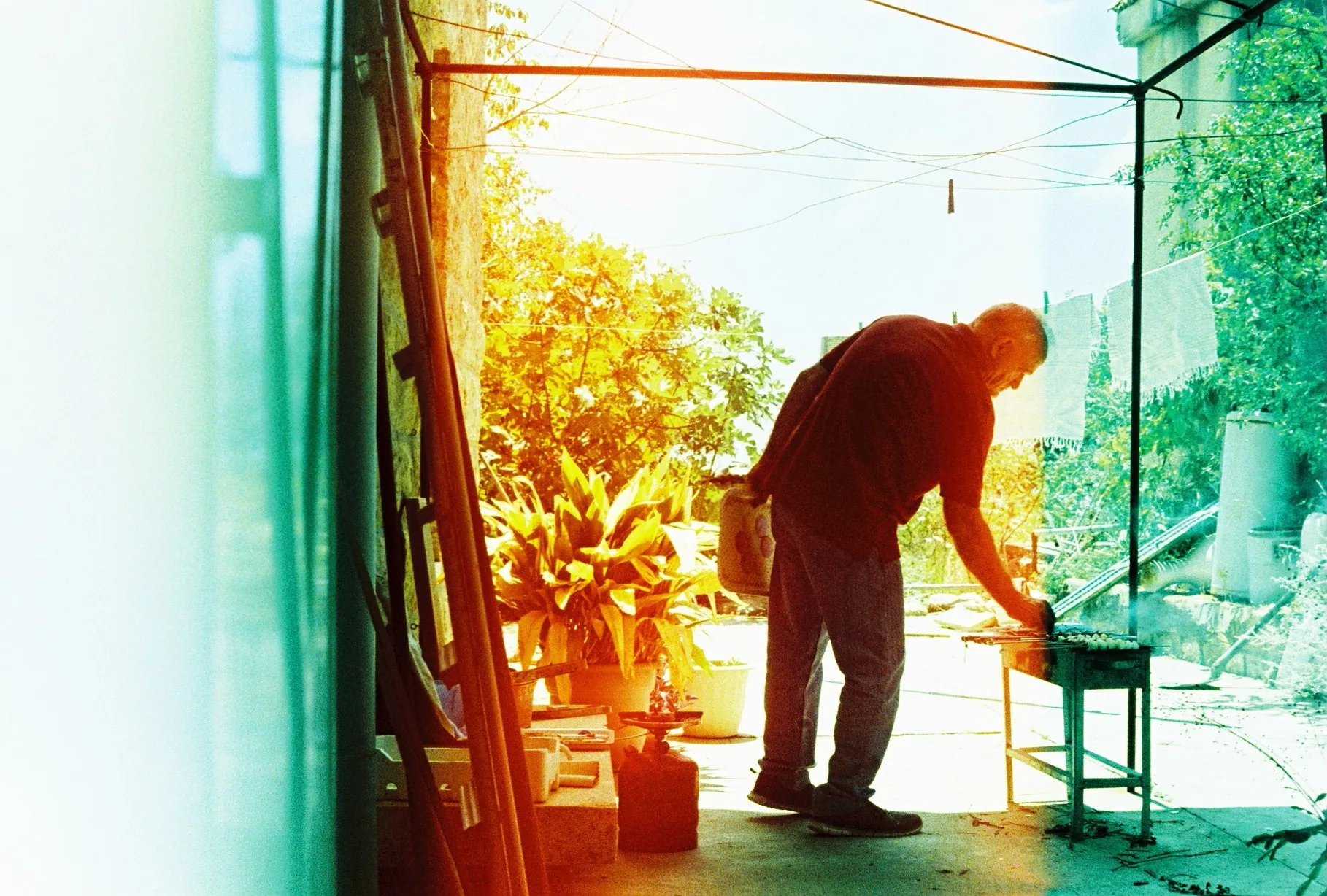 An older man leans over a low table on a cluttered terrace beside potted plants, bathed in warm orange light leak.