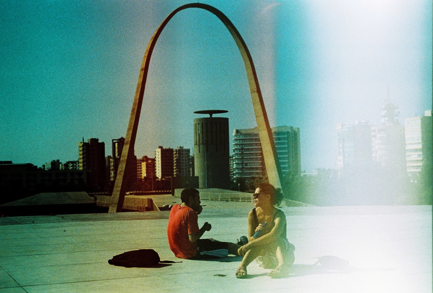 A man and a woman sit on bright pavement before a tall concrete arch with a city skyline behind them.
