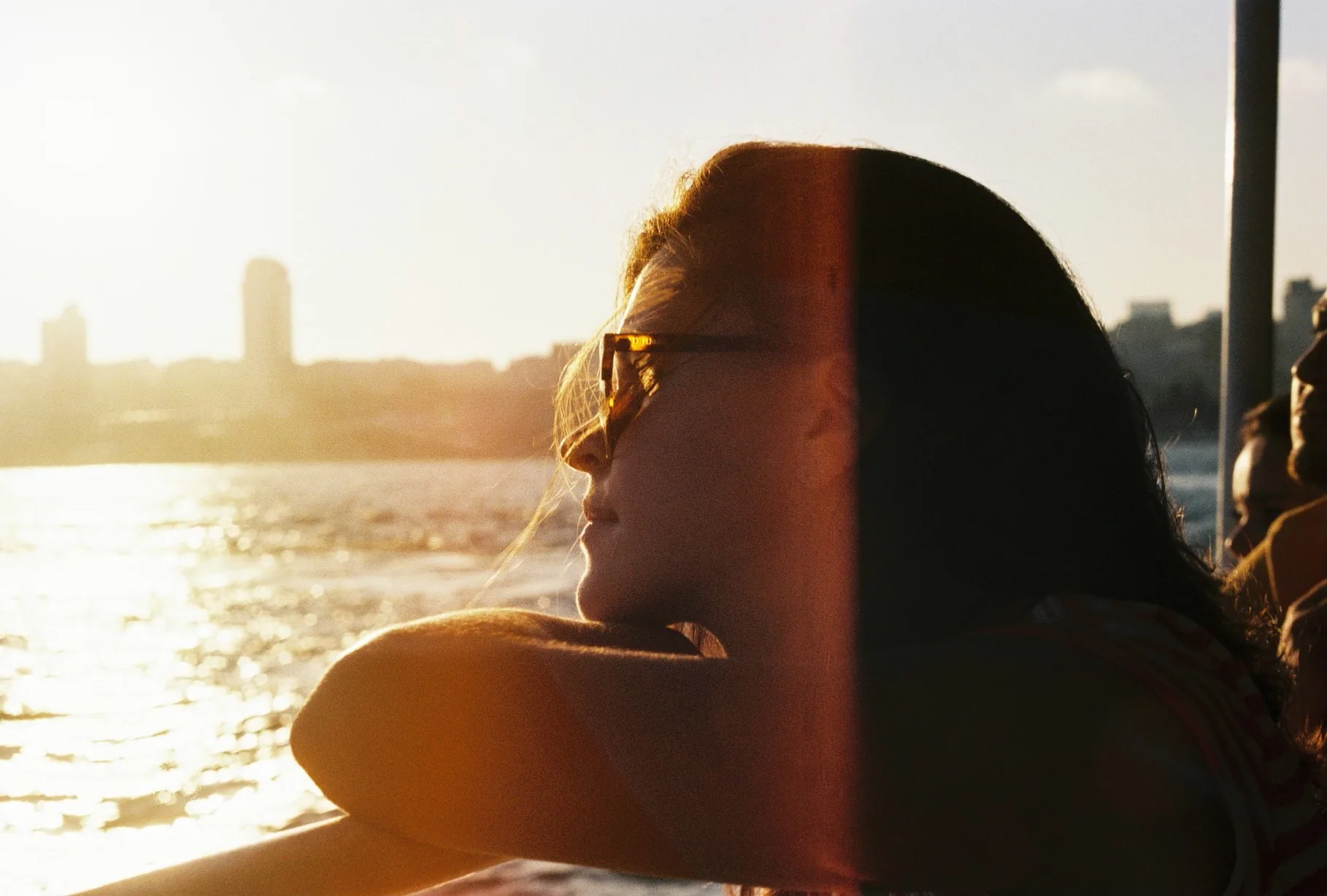 A woman in sunglasses leans back in profile against a sunlit railing, watching a hazy city skyline across the water at dusk.