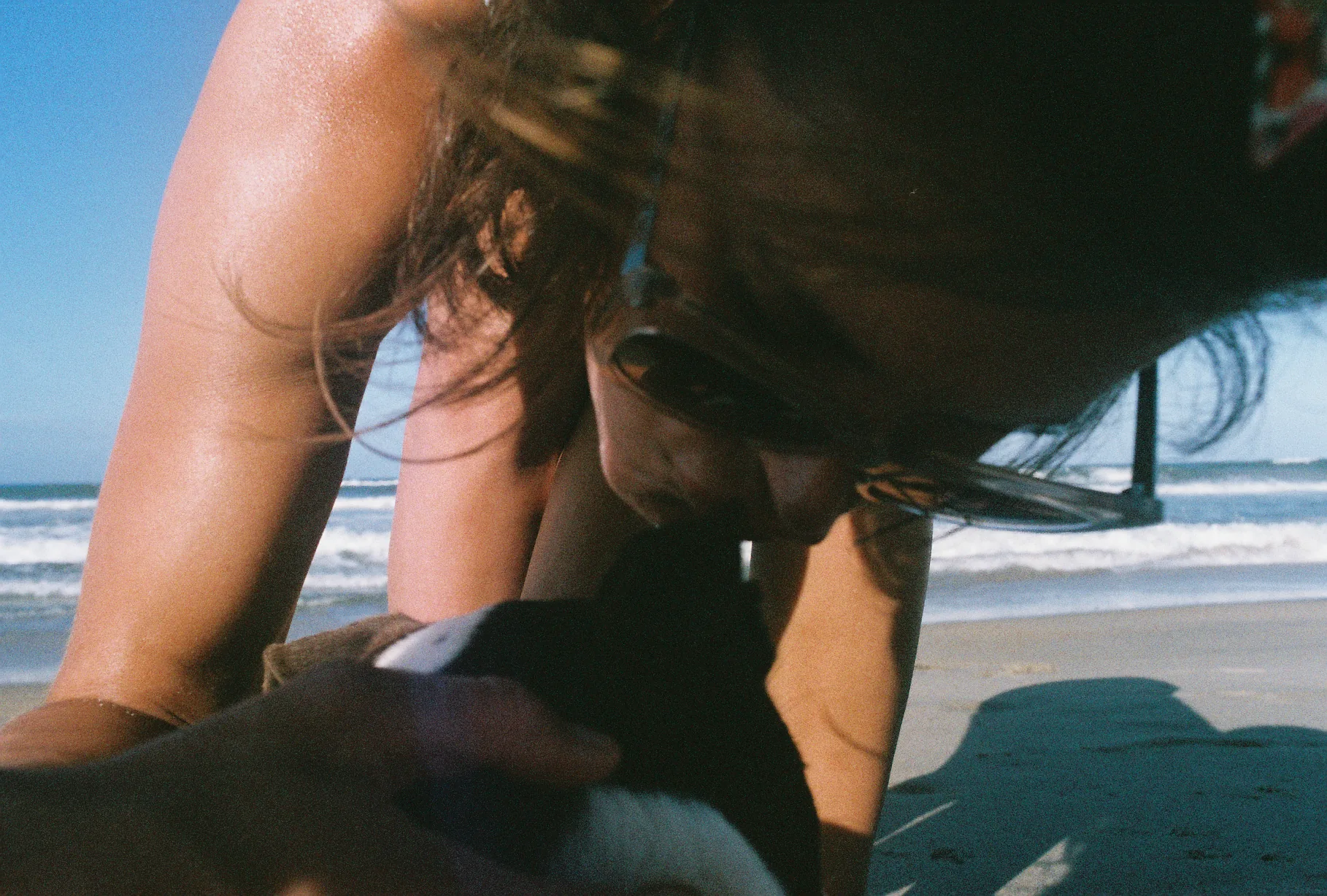 A woman in sunglasses bends down close to a black-and-white dog on a sandy beach with breaking waves behind them.