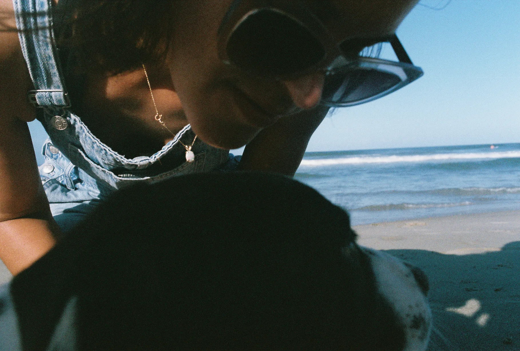 A woman in sunglasses and denim leans toward a black-and-white dog at the shoreline with waves rolling in behind.