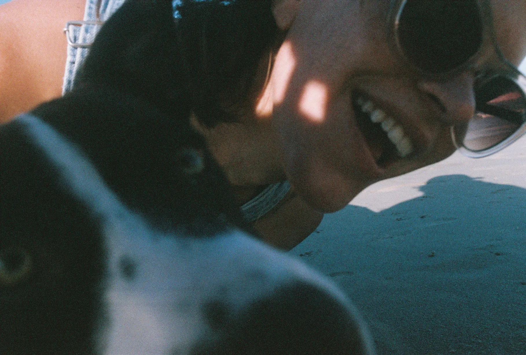 A smiling woman in sunglasses leans close to a black-and-white dog on the beach, both lit by warm low sunlight.