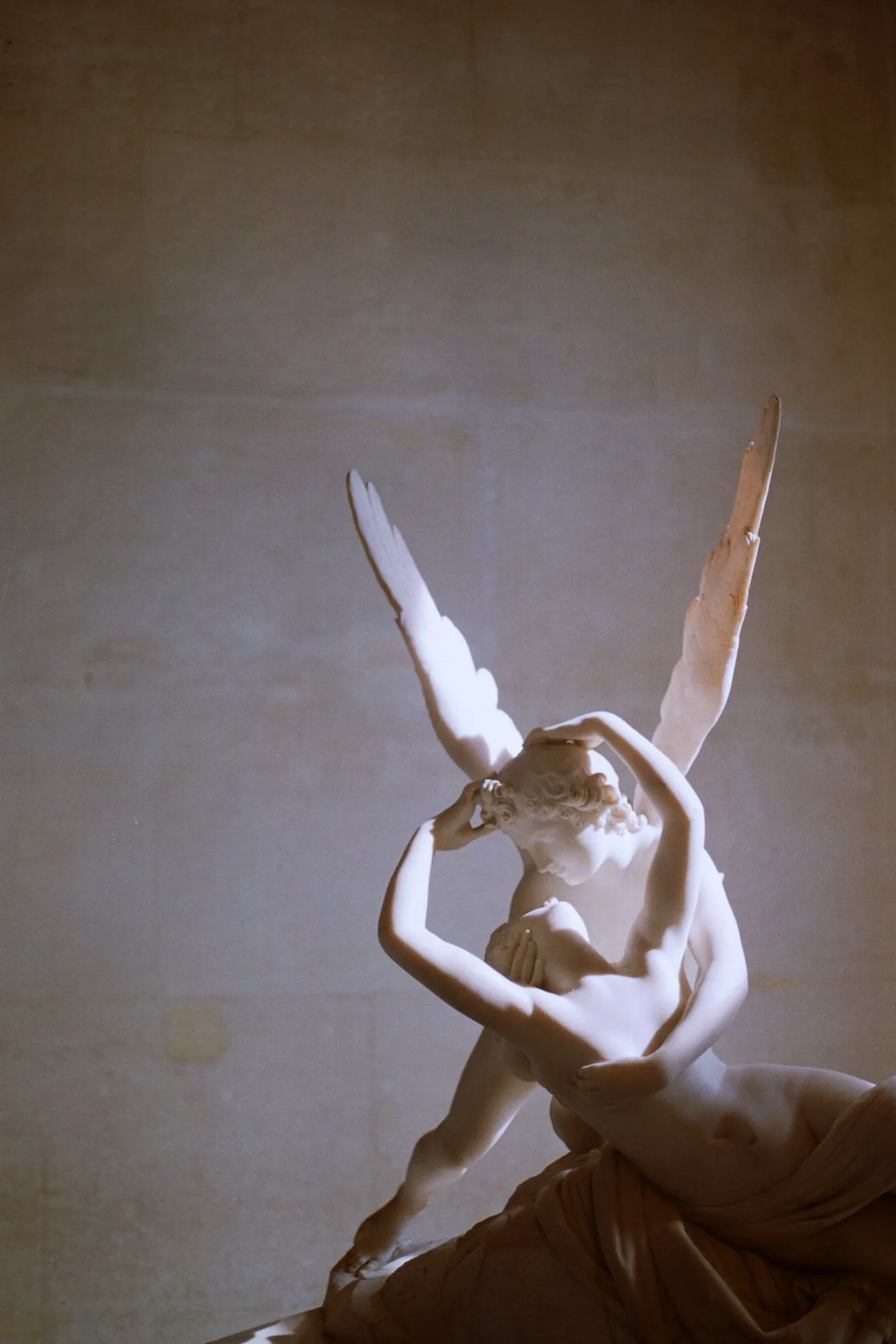 A white marble sculpture of a winged angelic figure cradling and kissing a reclining woman against a dim wall.
