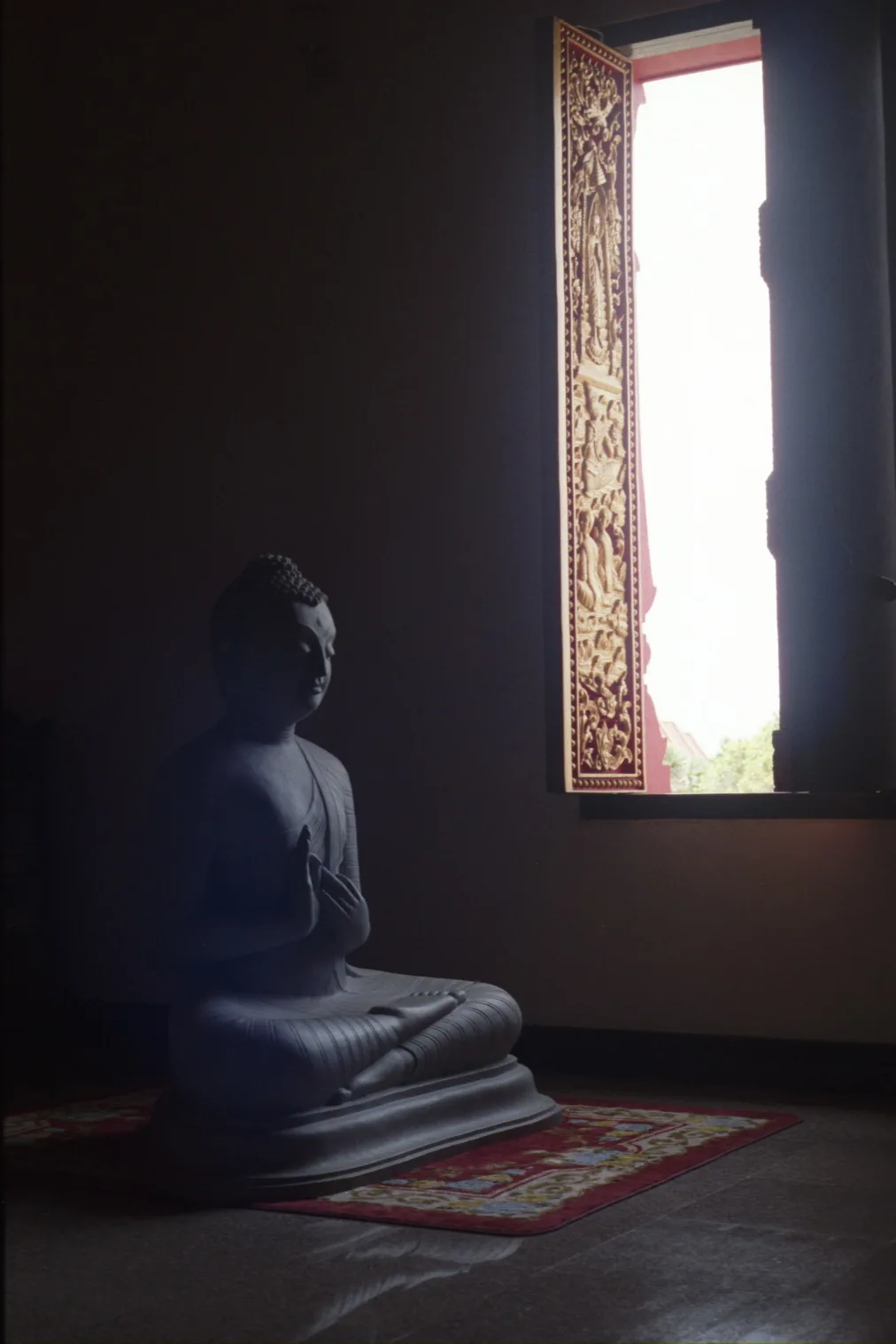 A dark stone seated Buddha rests on a red rug in a shadowed room beside an ornate carved window glowing with daylight.