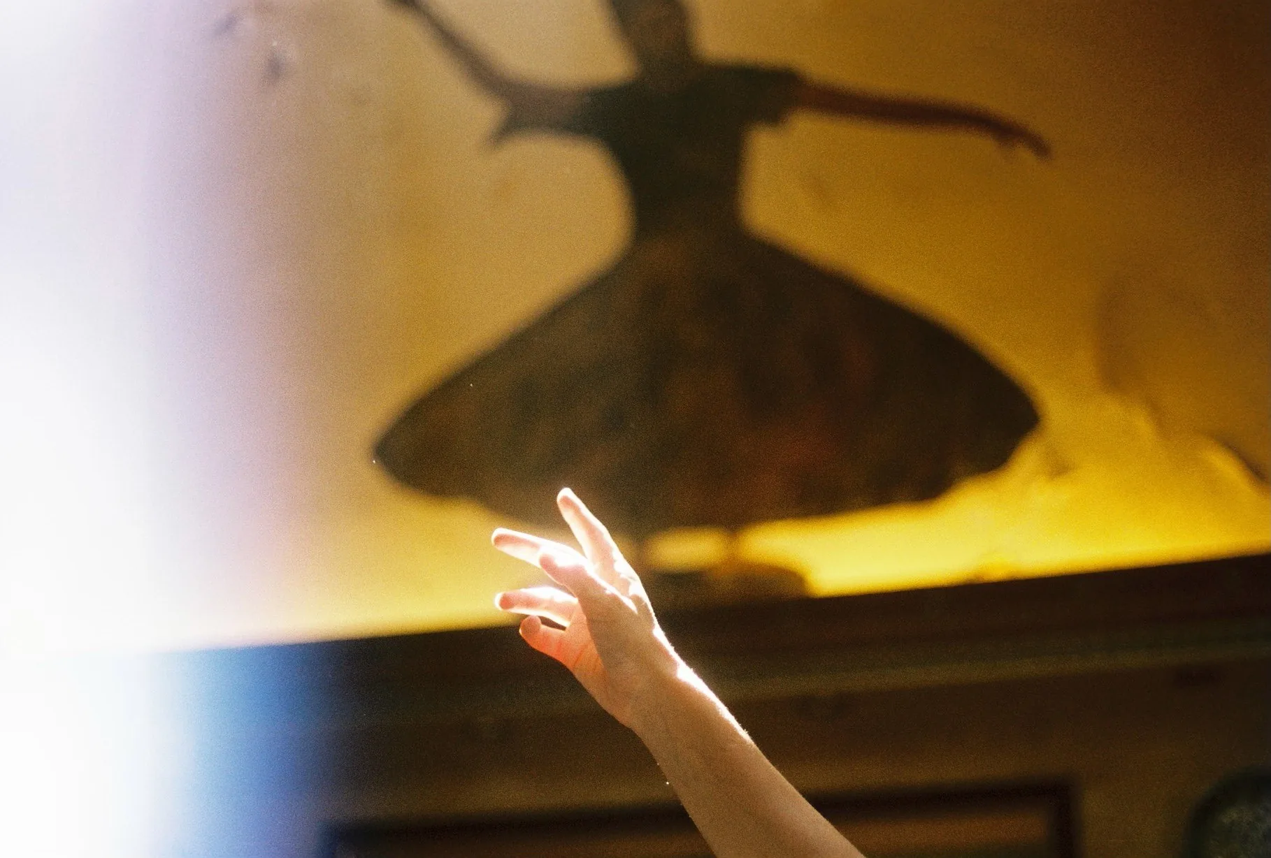 A backlit hand reaches upward toward the large shadow of a whirling dervish dancer cast on a warm golden wall.
