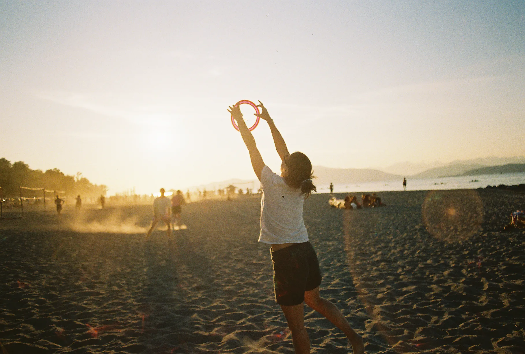 A person leaps with arms raised to catch a red flying disc on a hazy beach as the sun sets over distant mountains.