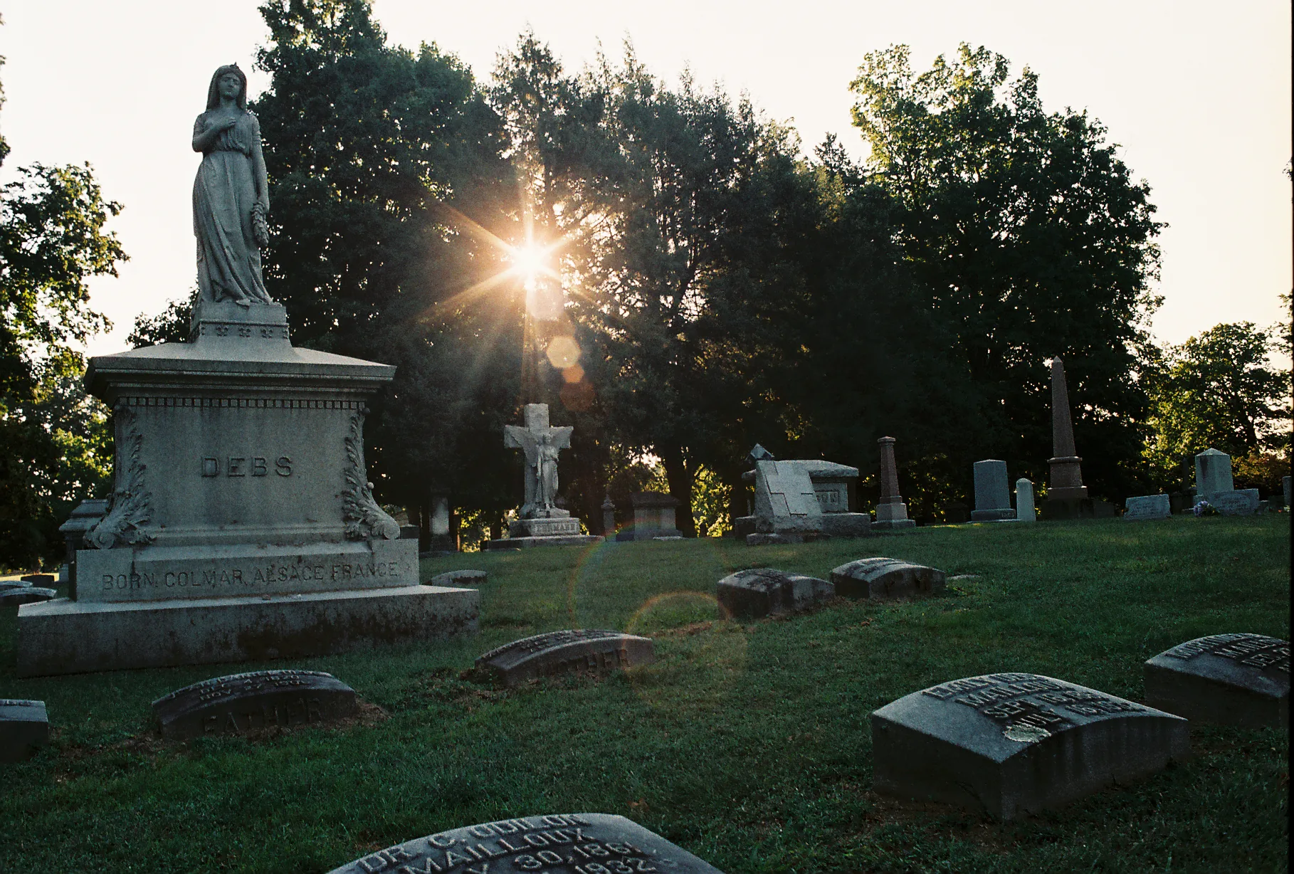 Sunlight bursts through trees behind a weathered statue and crucifix among scattered headstones in a grassy cemetery at dusk.