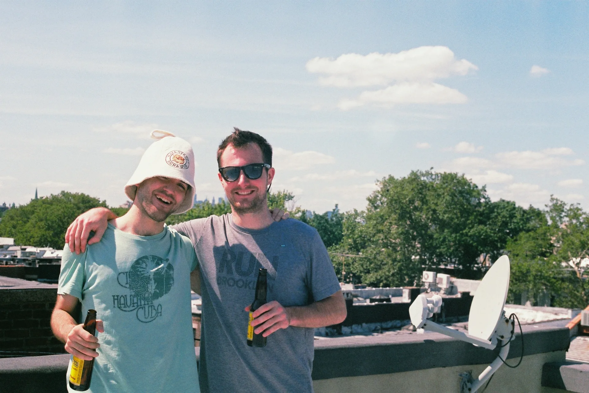 Two smiling men hold beer bottles on a rooftop with treetops and blue sky behind them on a bright sunny day.