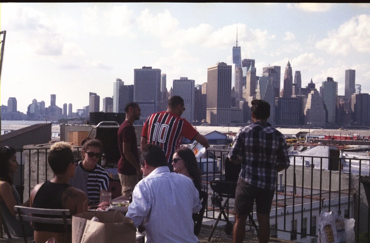 People mingle on a rooftop terrace overlooking a river and a dense downtown skyline of tall buildings under a blue sky.