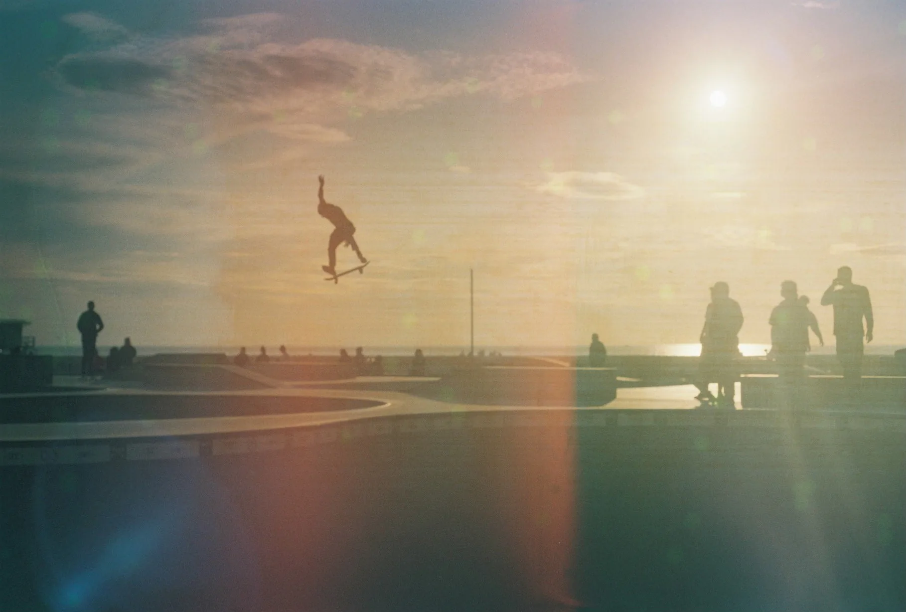 A skateboarder leaps mid-trick above a concrete skate bowl at sunset as silhouetted onlookers watch under a hazy sky with lens flare.
